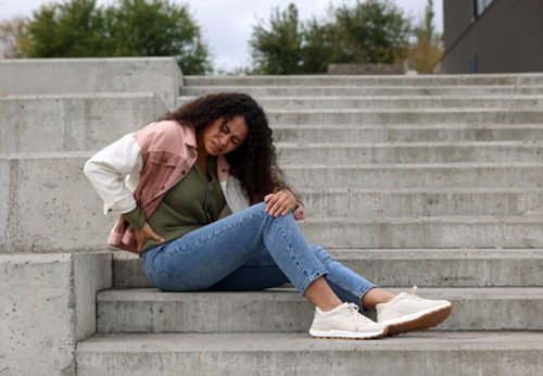 A young woman sitting on outdoor concrete steps, clutching her knee and back after an injury.
